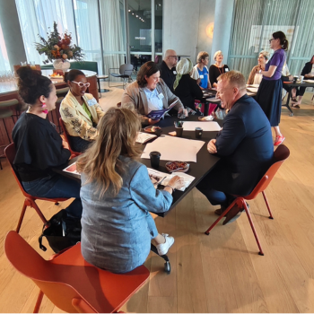 Group of people seated around a table in animated discussion during a workshop session in a bright room with large windows.